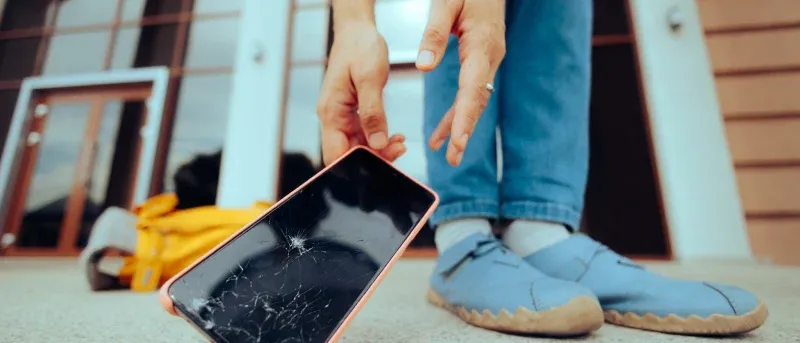 A person bends down to pick up a smartphone with a cracked screen from the ground.